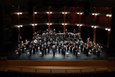 Ein großes Orchester steht auf der Bühne in einem Theater. Die Musiker sind in schwarzen Anzügen gekleidet und spielen verschiedene Instrumente. Der Dirigent führt die Aufführung. Die Beleuchtung ist gedämpft, was auf die musikalische Atmosphäre hinweist.